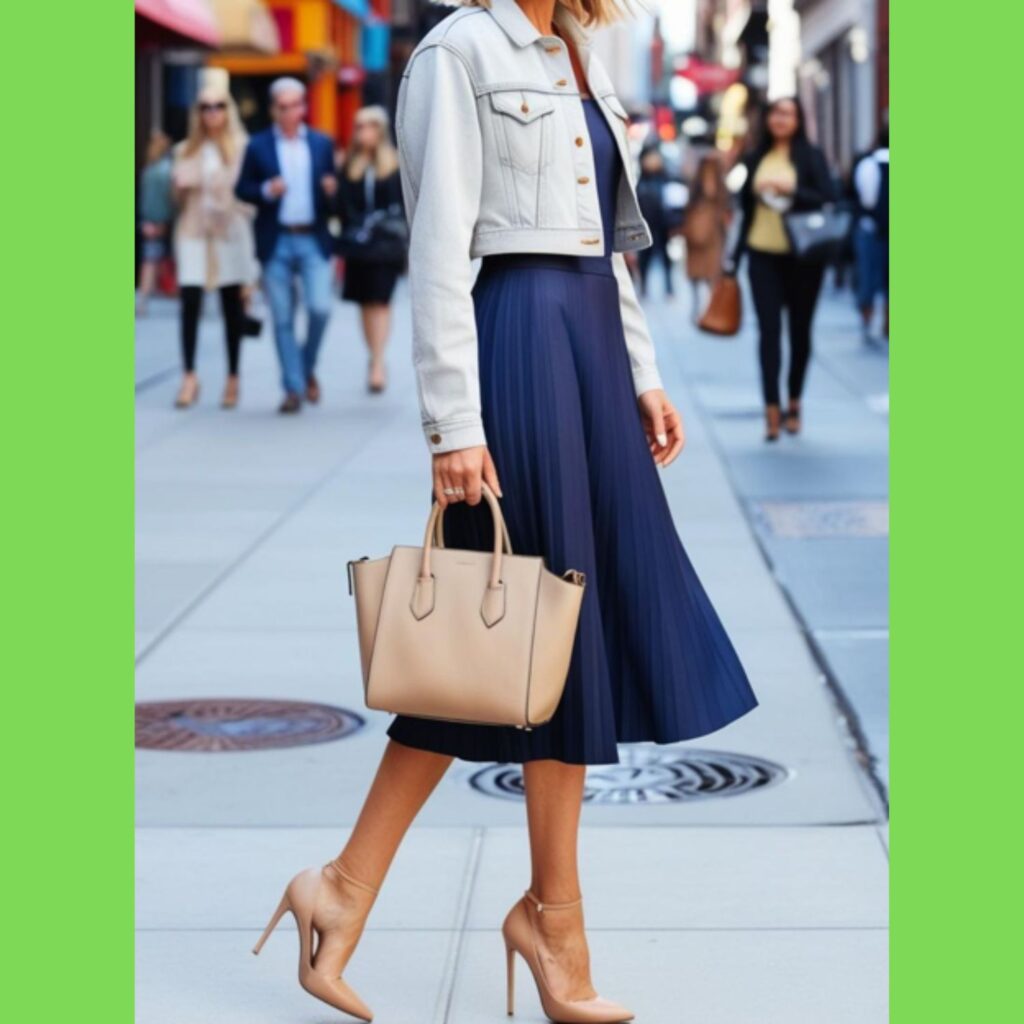 A beautiful woman in a cropped denim jacket with a pleated midi skirt and heels, carrying a handbag and posing on a city street.